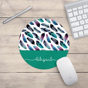 May include: Round teal mouse mat with a white background and a pattern of watercolour feathers in shades of blue, pink, and black. The name "Abigail" is written in white script on the teal section of the mouse mat.