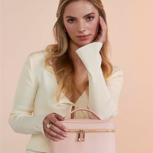 Pode incluir: Uma bolsa de cosm&eacute;ticos rosa claro com um z&iacute;per e uma al&ccedil;a douradas. A bolsa est&aacute; sentada no colo de uma pessoa que est&aacute; vestindo um su&eacute;ter branco.