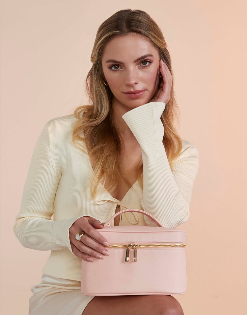 Pode incluir: Uma bolsa de cosm&eacute;ticos rosa claro com um z&iacute;per e uma al&ccedil;a douradas. A bolsa est&aacute; sentada no colo de uma pessoa que est&aacute; vestindo um su&eacute;ter branco.