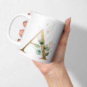 Op de afbeelding: Een witte keramische mok met een gouden letter A en groene aquarelbladeren. De mok heeft een handvat en wordt in een hand gehouden.