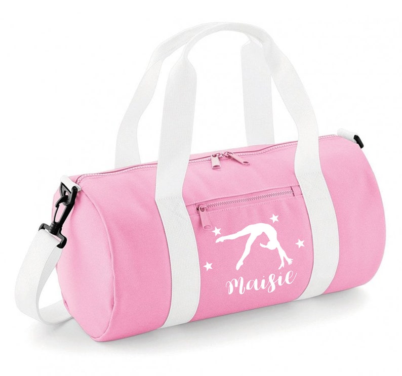 May include: Pink gym bag with white straps and a white silhouette of a gymnast doing a back handspring. The bag has white stars and the name "Maisie" printed on it.