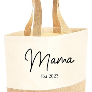 Może przedstawiać: Kremowa torba typu tote z napisem "Mama" czarnym skryptem i "Est 2023" wydrukowanym poniżej. D&oacute;ł i uchwyty są w naturalnym brązowym kolorze. Stylowy i praktyczny dodatek.