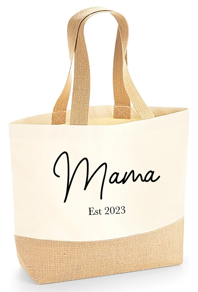 Może przedstawiać: Kremowa torba typu tote z napisem "Mama" czarnym skryptem i "Est 2023" wydrukowanym poniżej. D&oacute;ł i uchwyty są w naturalnym brązowym kolorze. Stylowy i praktyczny dodatek.