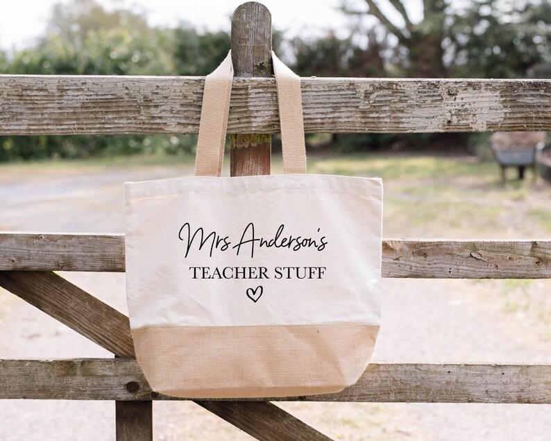 May include: A beige canvas tote bag with brown handles. The bag has the text "Mrs. Anderson's Teacher Stuff" printed in black with a small heart symbol.