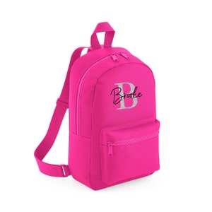 Puede incluir: Una mochila de color rosa brillante con un bolsillo frontal con cremallera. La mochila tiene una letra "B" de purpurina plateada y el nombre "Brooke" bordado en la parte delantera.