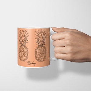 Pode incluir: Uma caneca de cerâmica branca com fundo pêssego e desenhos de linha preta de dois abacaxis. A caneca tem uma inscrição preta que diz "Bailey".