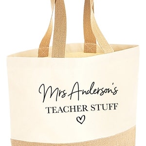 May include: A beige canvas tote bag with brown jute handles and a brown jute bottom. The bag has black text that says "Mrs. Anderson's Teacher Stuff" with a heart symbol.