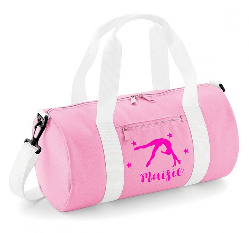 May include: Pink gym bag with white straps and a zipper closure. The bag has a pink gymnast silhouette with stars and the name "Maisie" printed on it.