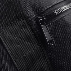May include: Close-up of a black zipper on a black fabric bag. The zipper is metal and has a black pull tab.