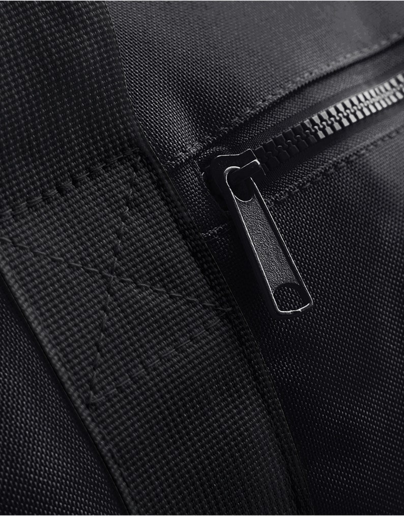 May include: Close-up of a black zipper on a black fabric bag. The zipper is metal and has a black pull tab.