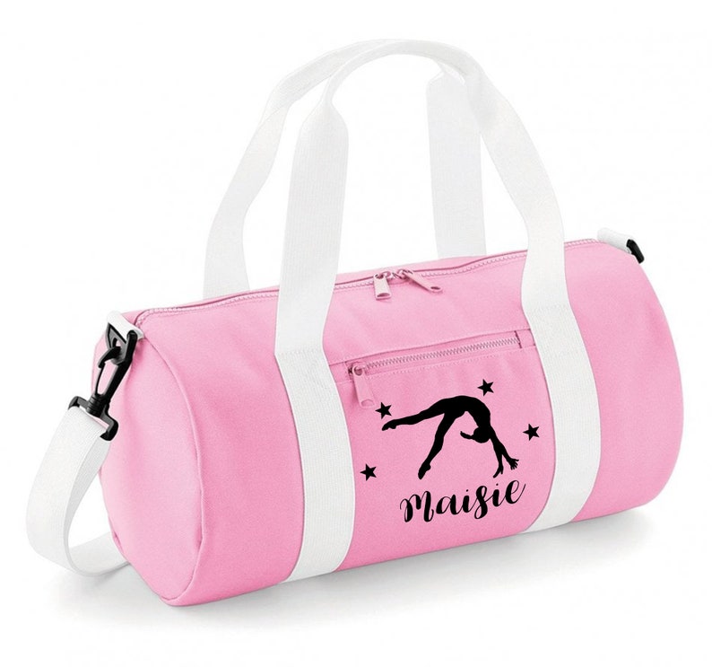 May include: Pink gym bag with white straps and a black silhouette of a gymnast doing a back handspring. The bag has a zippered pocket and the name "Maisie" is printed on the front.