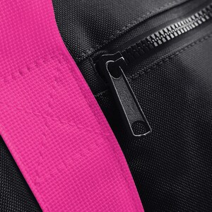 May include: Close-up of a black zipper on a black bag with a bright pink strap.