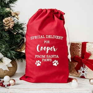 Może przedstawiać: Czerwony materiałowy worek na prezenty z białym tekstem, który brzmi "Special Delivery for Cooper From Santa Paws" z dwoma białymi odciskami łap.