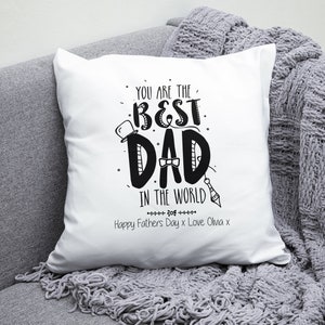 Op de afbeelding: Een wit kussen met zwarte tekst die "You are the best Dad in the world" luidt, met een strik en een hoge hoed. De tekst "Happy Father's Day x Love Olivia x" is ook inbegrepen.