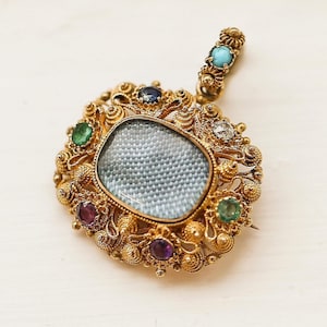 Puede incluir: Un colgante de oro con un delicado diseño de filigrana y un pequeño camafeo de forma ovalada. El camafeo está rodeado de piedras preciosas de colores, incluyendo turquesa, esmeralda y amatista.