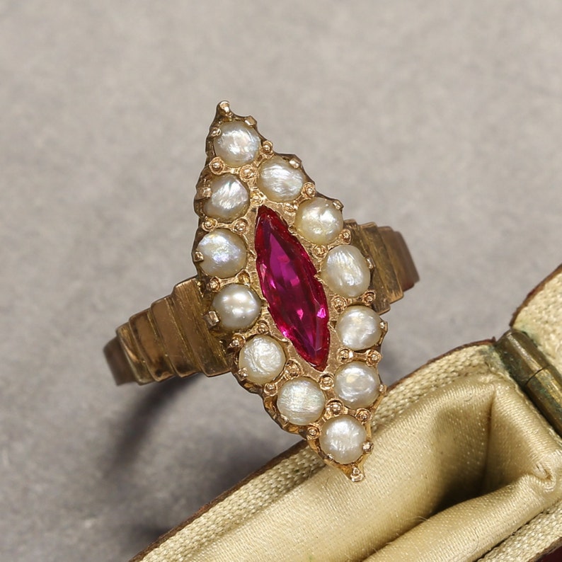 May include: A gold ring with a red gemstone surrounded by white pearls. The ring is in the shape of a teardrop.