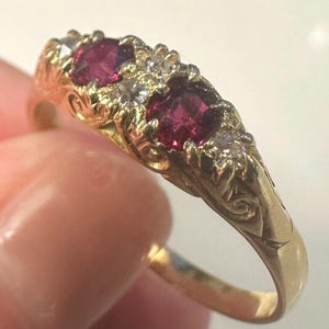 Puede incluir: Un anillo de oro con tres piedras preciosas ovaladas de color rojo intenso, posiblemente rubíes, acentuadas por pequeñas piedras transparentes. El anillo tiene un diseño floral ornamentado en la banda y alrededor de las piedras preciosas. El anillo está sostenido por una mano.
