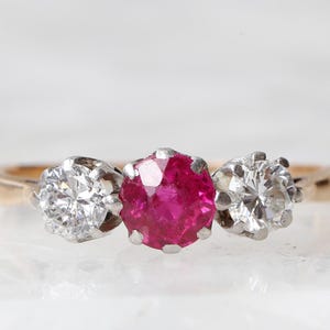 May include: A three-stone ring featuring a central round-cut vibrant red ruby flanked by two round-cut clear white diamonds. The stones are set in a silver-toned metal setting, which is mounted on a polished yellow gold band.