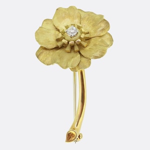 Könnte beinhalten: Eine goldene Blumenbrosche mit einer texturierten, matten Oberfläche auf den Blütenblättern und einem einzelnen, runden, klaren Edelstein in der Mitte. Die Blume ist an einem glatten, geschwungenen Goldstiel befestigt, der nach unten führt.