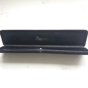 May include: An open, black velvet jewelry box with the brand name "DeLaneau" in silver lettering. The box is rectangular and has a small, round clasp. The interior is also black.