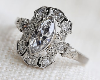Puede incluir: Un anillo de plata con un gran diamante de forma marquesa rodeado de diamantes más pequeños. El anillo tiene un diseño delicado e intrincado.