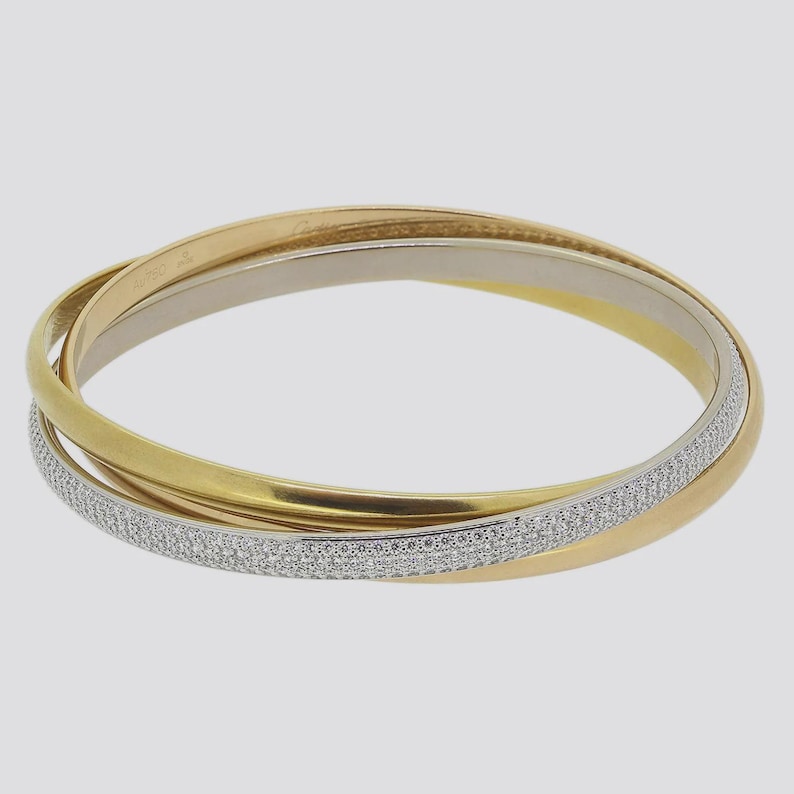 May include: A gold, silver, and white bangle bracelet with a diamond-encrusted band. The bracelet is made of three interlocking bands, each with a different finish. The diamond-encrusted band is the bottom band and is the most prominent feature of the bracelet.