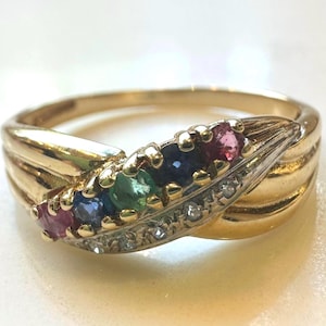 Könnte beinhalten: Ein goldener Ring mit geschwungenem Design, mit einer Reihe farbiger Edelsteine, darunter Rubine, Saphire, Smaragde und Diamantakzente. Der Ring hat eine polierte Oberfläche und einen klassischen, eleganten Stil.