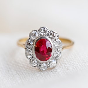 Puede incluir: Anillo de oro con un rubí central ovalado, rodeado por un halo de pequeños diamantes. El engaste combina oro y plata, con una banda delgada. El anillo se muestra sobre un fondo blanco suave, resaltando su elegante diseño.