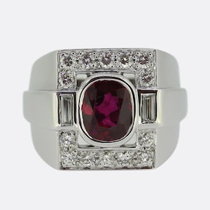 May include: A white gold ring featuring a large oval ruby center stone. The ruby is surrounded by a rectangular frame of small, round diamonds. Two rectangular diamonds flank the ruby on each side.