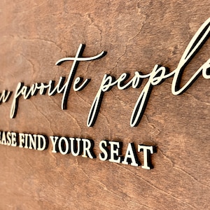 May include: A wooden sign with the text "Our favorite people, please find your seat" in gold lettering.