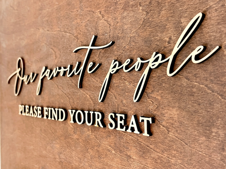May include: A wooden sign with the text "Our favorite people, please find your seat" in gold lettering.