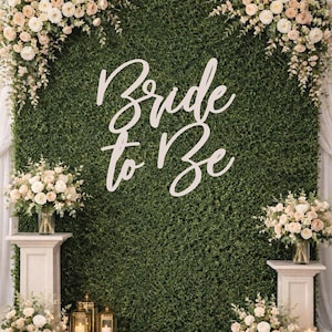 May include: A green hedge wall backdrop with the words "Bride to Be" in white script. The backdrop is decorated with floral arrangements and candles, creating a romantic setting for a wedding or bridal shower.