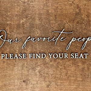 May include: A wooden sign with the text "Our favorite people, please find your seat" in white lettering.