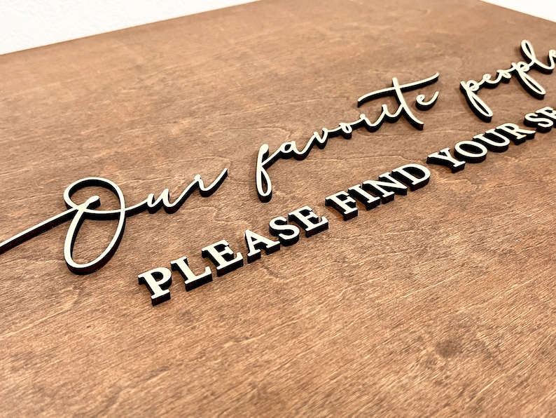May include: A wooden sign with the text "Our favorite people please find your seat" in black lettering.