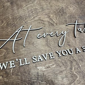 May include: Wooden sign with the words "At every Table, We'll save you a seat" in white, elegant lettering. The sign is made of a dark brown wood with visible grain patterns, creating a rustic aesthetic.