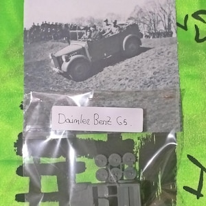 Può includere: Fotografia in bianco e nero di un veicolo militare Daimler Benz G5. L'immagine mostra il veicolo su una strada sterrata con soldati sullo sfondo. Sotto la foto ci sono parti di un modello e un'etichetta che dice "Daimler Benz G5."