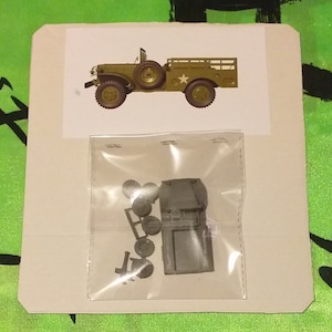 Puede incluir: Una bolsa de plástico que contiene un kit de maqueta de un camión militar. El kit incluye la carrocería del camión, las ruedas y otras piezas. El camión está pintado con un patrón de camuflaje verde.