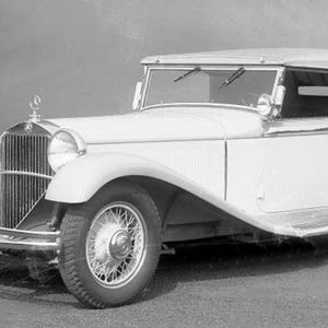 Può includere: Un'auto d'epoca cabriolet color crema con capote. L'auto ha un'imponente griglia anteriore, grandi fari e ruote a raggi. La targa recita IIIA-21905. L'auto è parcheggiata su una superficie pavimentata.