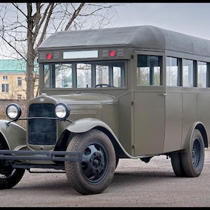 Pode incluir: Um autocarro vintage verde-azeitona com carroçaria quadrada, tejadilho arredondado e pneus pretos grandes. O veículo apresenta uma grelha frontal proeminente, dois faróis circulares, um para-choques dianteiro preto e janelas laterais retangulares.