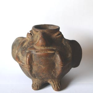 May include: A small, brown, ceramic vessel with a rounded body and a narrow opening. The vessel has three small feet and a textured surface. The design includes four rounded protrusions and a textured rim.