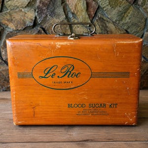 May include: A rectangular wooden box with a metal handle and latch. The front features a green oval logo with the text Le Roc TRADE MARK and the text BLOOD SUGAR KIT MANUFACTURED BY LE ROC LABORATORIES MIDDLEBORO, MASSACHUSETTS printed in black.