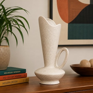May include: A cream-colored ceramic vase with a textured, granular surface. The vase features a wide, rounded base, a tall, flared neck with an asymmetrical opening, and a single circular handle attached to the side of the neck and body.