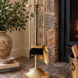 May include: A gold-toned metal fireplace tool set featuring a stand with a shell-shaped base, a poker, a shovel, and a brush with black bristles. Each tool handle is topped with a decorative shell finial, and the set is organized on a central vertical rod.