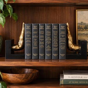 May include: A set of two black L-shaped bookends featuring gold-colored human hand sculptures, positioned on either side of six black Reader's Digest books with gold lettering on the spines, arranged upright on a wooden shelf.