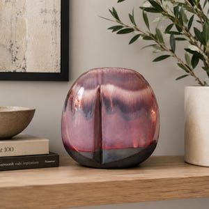 May include: A rounded ceramic vase featuring a reactive glaze with blended tones of deep burgundy, soft mauve, and dark charcoal. The surface has a smooth, glossy finish with a vertical indentation running down the center of the front face.