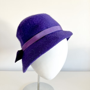 Puede incluir: Un sombrero de fieltro morado con una cinta morada a juego. El sombrero está en un expositor blanco.