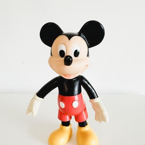 Pode incluir: Uma figura de plástico do Mickey Mouse em preto e branco com calções vermelhos e sapatos amarelos. A figura está em pé sobre um fundo branco.