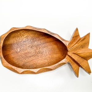 May include: A wooden pineapple-shaped bowl with a smooth, brown finish. The bowl is carved with a detailed pineapple design and has a wide, shallow opening.
