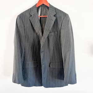Op de afbeelding: Een zwart-wit gestreept blazer hangend aan een rode houten hanger. De blazer heeft een enkele knoopsluiting en twee zakken.