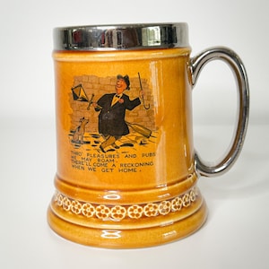 May include: A brown ceramic beer stein with a silver lid and handle. It features a cartoon illustration of a man with an umbrella and a dog. The text on the stein reads: "Thro' pleasures and pubs we may roam. There'll come a reckoning when we get home."